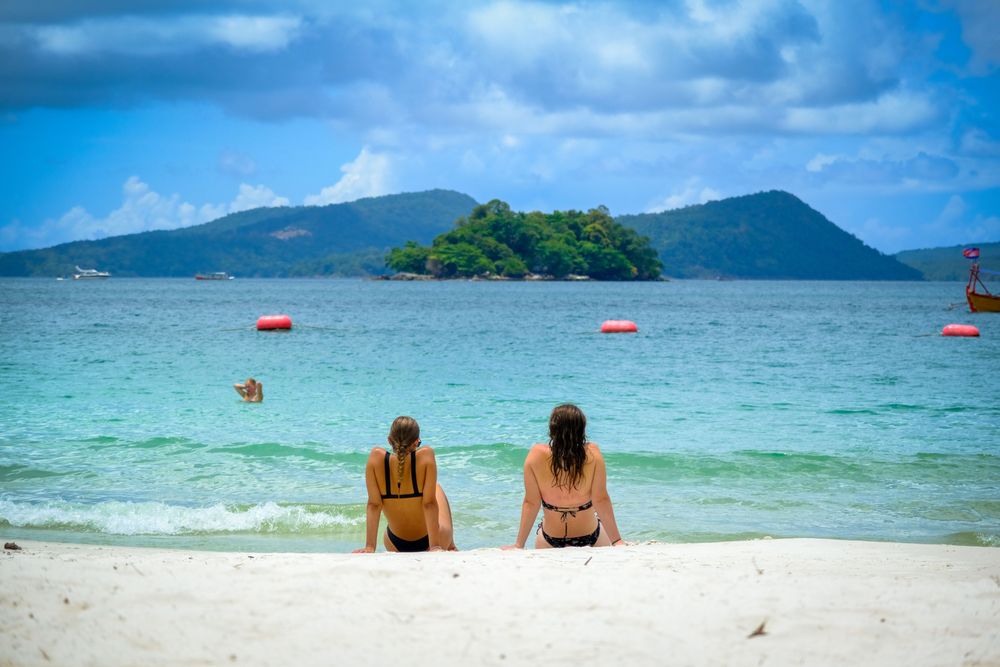 female friends sitting on a pristine white sand beach in Phuket looking out at a tropical green island across clear turquoise water under a blue sky matching the file ssa beach girls.jpg
