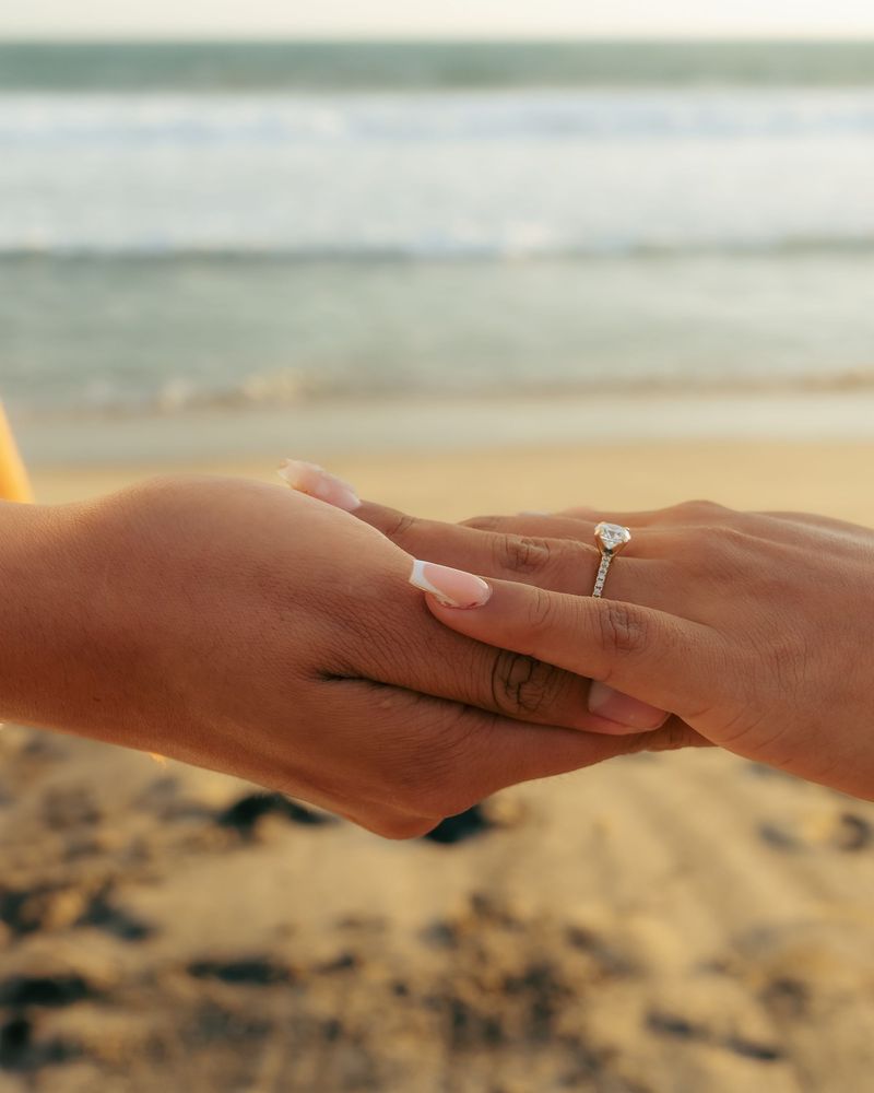 Close up view of a newly engaged couple holding hands on a sandy beach at sunset with a gold diamond engagement ring visible on her finger against a blurred ocean wave