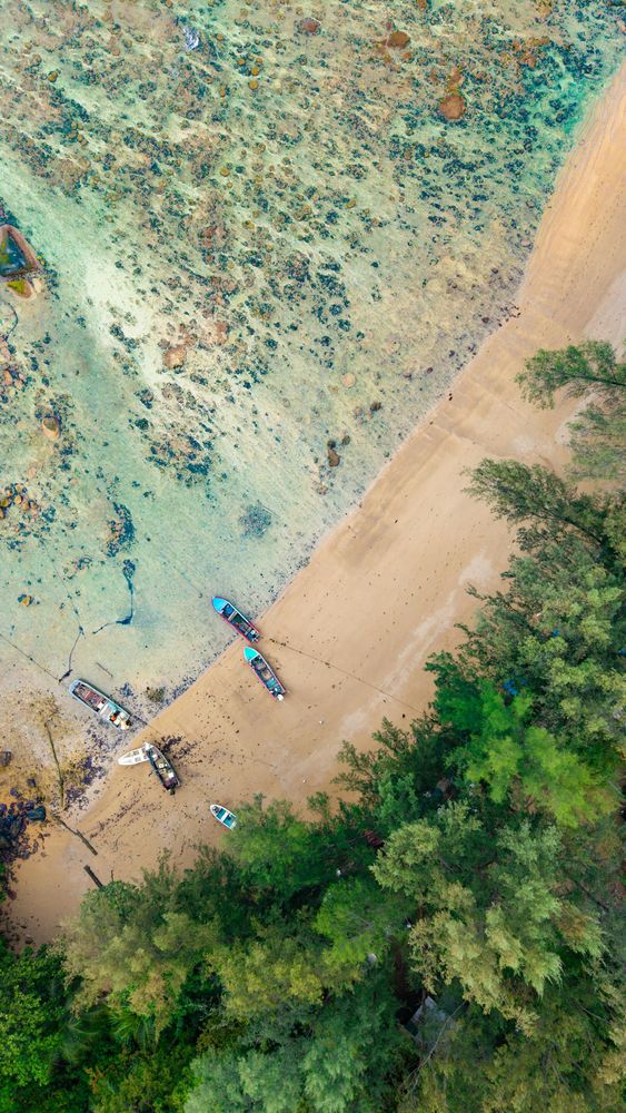 Aerial top-down view of a turquoise coral reef and sandy beach in southern Phuket with longtail boats moored at the shore and dense tropical trees — the kind of breathtaking shot only possible from a Silk Sky Air helicopter
