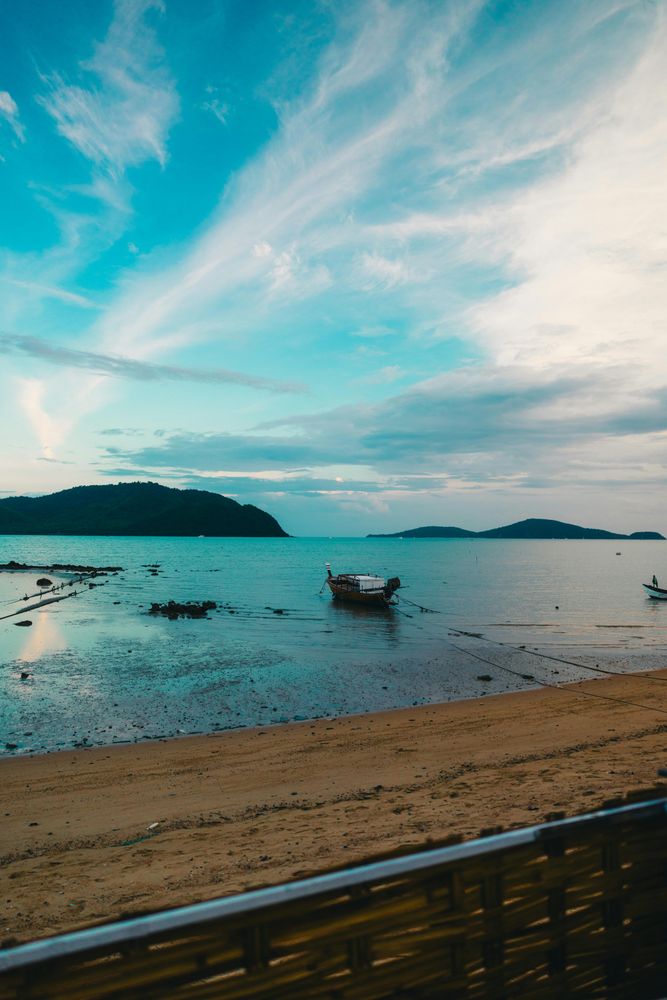 Longtail boat moored at calm Rawai Beach in southern Phuket at dusk, with turquoise water, sandy shore and distant islands — the destination reached in minutes via Silk Sky Air helicopter tour