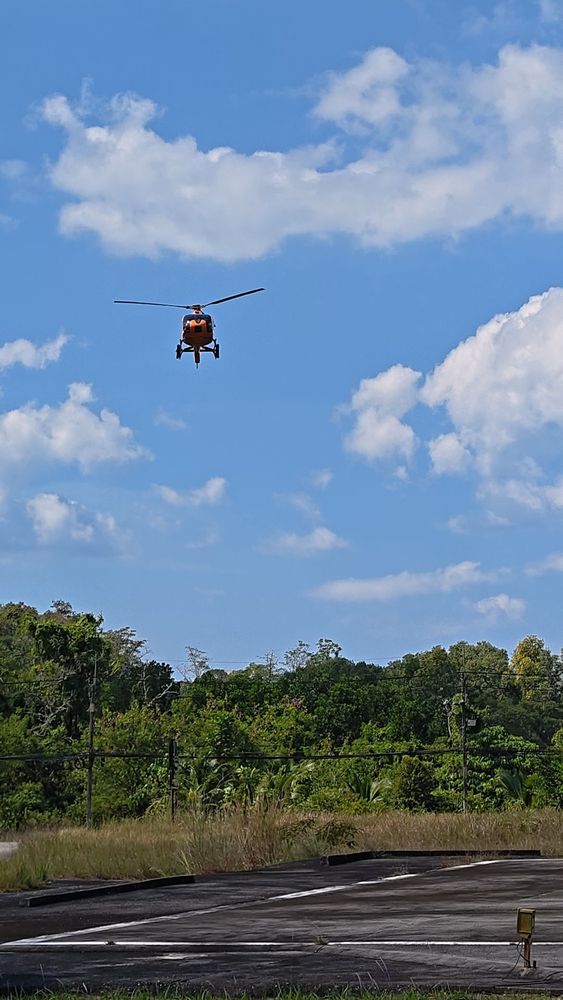 A Silk Sky Air orange helicopter lifting off into a clear blue sky above the helipad in Phuket, surrounded by lush tropical greenery
