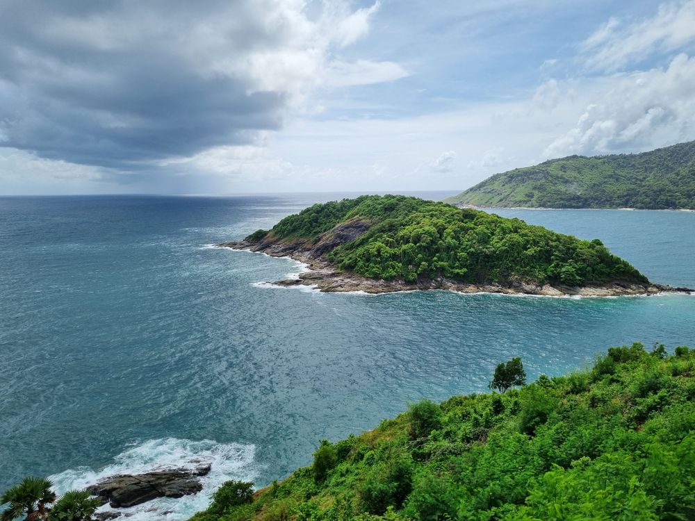A scenic wide panoramic view of a lush green tropical island surrounded by deep blue ocean waters under a dramatic cloudy sky in Phuket